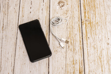 A black smartphone with a carbon fiber case on a vintage white wooden table and a white charging cable