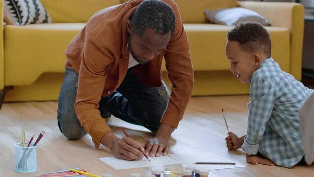 Cute little african american boy and his daddy drawing pictures together, creating on floor at home, tracking shot