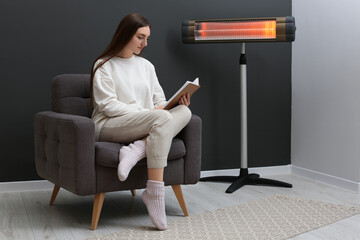 Young woman reading book on armchair near electric infrared heater at home. Space for text