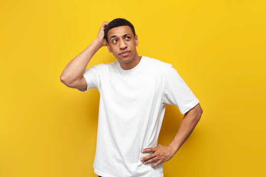 young puzzled african american guy in white t-shirt sketches his head with his hand on yellow isolated background
