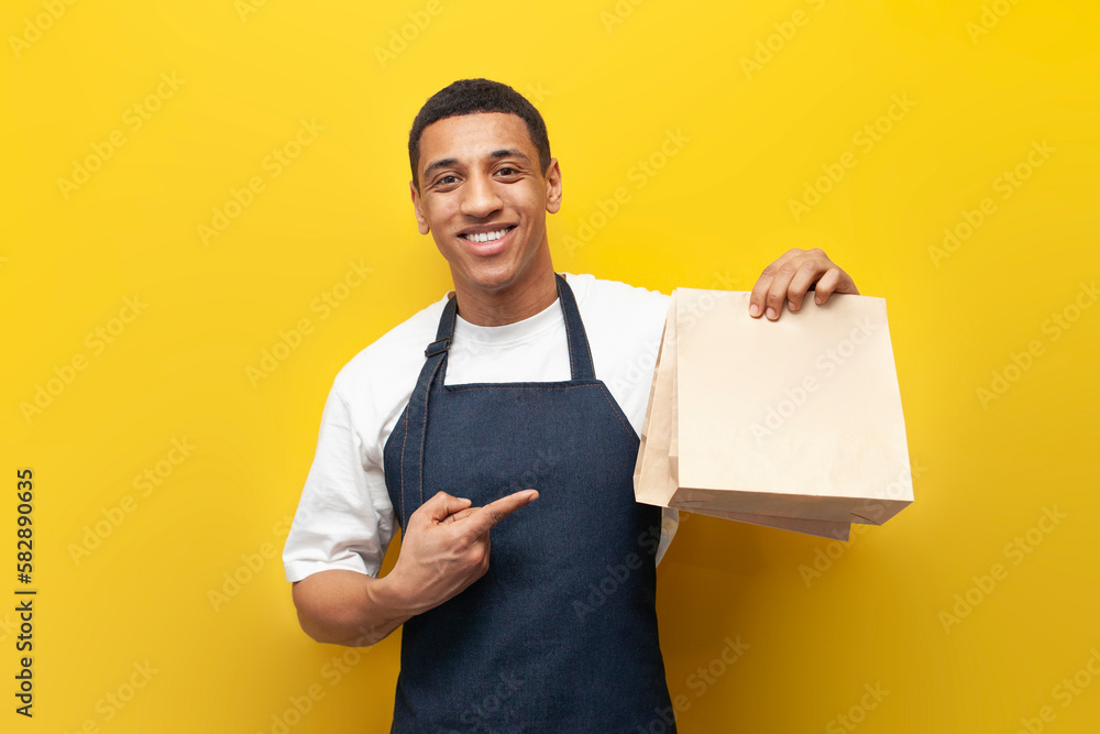 Wall mural young African American barista guy in uniform holds packages of food on yellow background, delivery service