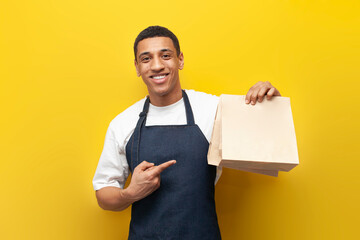 young African American barista guy in uniform holds packages of food on yellow background, delivery service