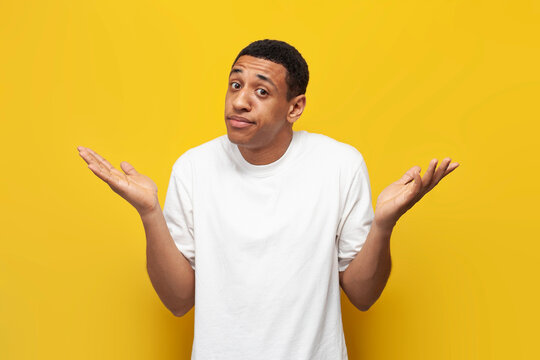 young insecure African American guy in white T-shirt shrugs his shoulders, confused man is not sure and does not know