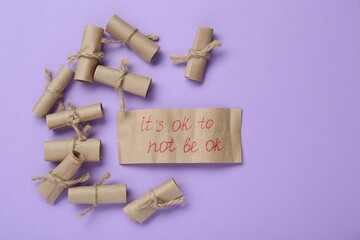 Card with phrase It`s Ok to Not Be Ok and rolled pieces of paper on violet background, flat lay....