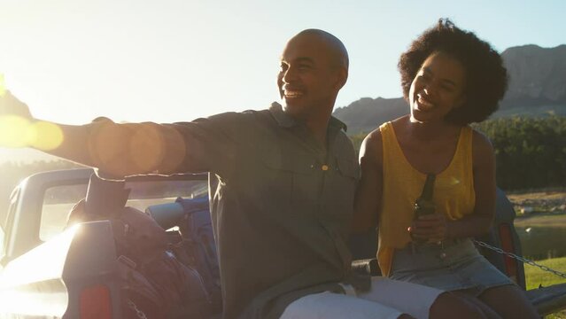 Couple with backpacks sitting on tailgate of pick up truck on road trip in countryside drinking beer and doing cheers  - shot in slow motion