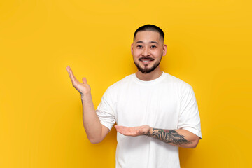 adult tattooed asian man in white t-shirt shows his hands to the side on yellow isolated background...