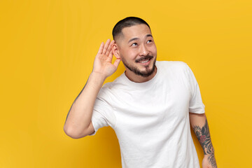 adult tattooed asian man in white t-shirt eavesdrops on yellow isolated background and smiles