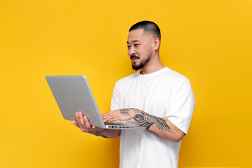 adult tattooed asian man in white t-shirt uses laptop on yellow isolated background, unshaven korean guy