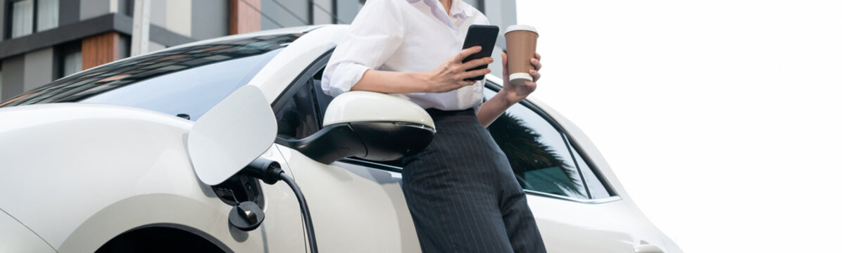 Focus businessman using phone, leaning on electric vehicle, holding coffee with blurred city residential condo buildings in background as progressive lifestyle by renewable and sustainable EV car.