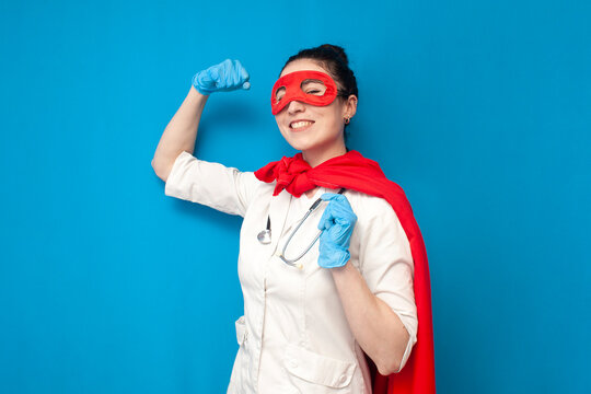 cheerful young girl doctor in uniform in superman costume on blue background, female nurse superhero shows strength