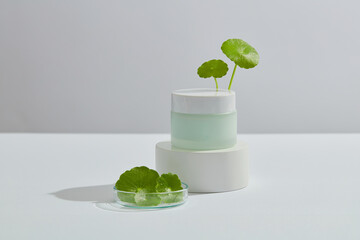 Front view of cream jar without label on podium and gotu kola leaves on petri dish on white...