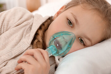 Little girl using nebulizer for inhalation on bed indoors, closeup