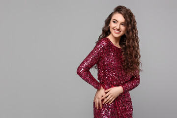Beautiful young woman with long curly brown hair in pink sequin dress on grey background, space for text