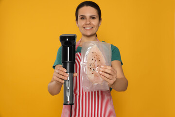 Beautiful young woman holding sous vide cooker and meat in vacuum pack against orange background,...