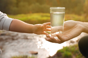 Mother giving her daughter glass of fresh water near stream on sunny day, closeup - obrazy, fototapety, plakaty