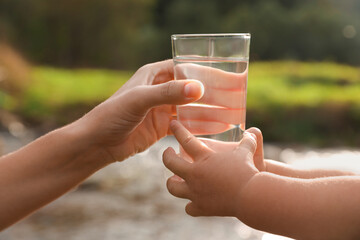 Mother giving her daughter glass of fresh water near stream on sunny day, closeup