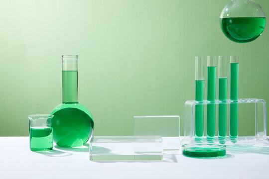 Test tubes, petri dish, beaker and flat-bottom florence flask containing green fluid. Empty transparent podium in rectangle shape for product mockup