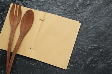 Blank recipe book and wooden utensils on black textured table, flat lay. Space for text
