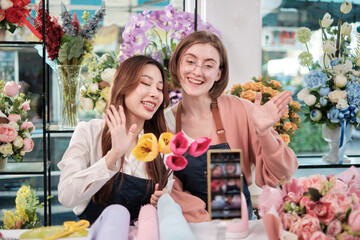 E-commerce business, two young female florist workers demonstrate and show floral arrangements via online live streaming with smartphone application in bright flower shop, a beautiful blossoms store.
