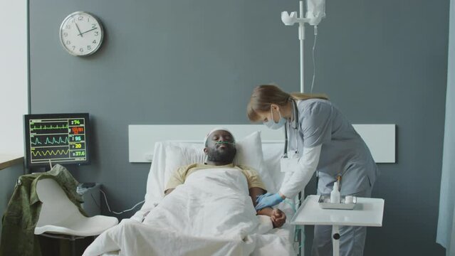 Medium long shot of nurse adding medication to IV bag for African American patient in military hospital