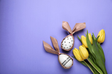 Easter bunnies made of craft paper and eggs near beautiful tulips on purple background, flat lay. Space for text