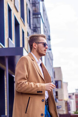 A young guy in sunglasses and a beige stylish coat smokes on the street in the city. Bad habit