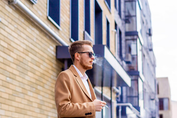 A young guy in sunglasses and a beige stylish coat smokes on the street in the city. Bad habit