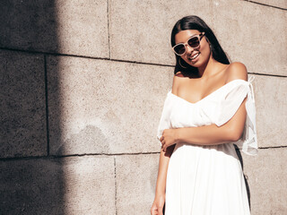Young beautiful smiling hipster woman in white dress. Sexy carefree model posing on the street background at sunset. Positive female outdoors at warm sunny day. Cheerful and happy. In sunglasses