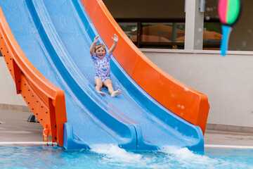 Little preschool girl sliding on a children slide in outdoor swimming pool in hotel resort. Child learning to swim in outdoor pool, splashing with water, laughing and having fun. Family vacations.