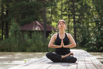 Fototapeta na wymiar A woman leading a healthy lifestyle and practicing yoga is engaged in meditation sitting in a lotus position on a wooden path on a sunny summer morning in the park