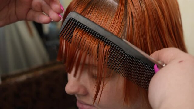 The hairdresser cuts the bangs of a red-haired European girl with her eyes closed with scissors in a barbershop. Close-up side view