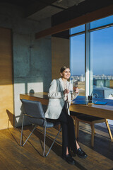 Office work, attractive young female manager working in office, standing in bright workspace using modern laptop computer, young woman drinking delicious coffee