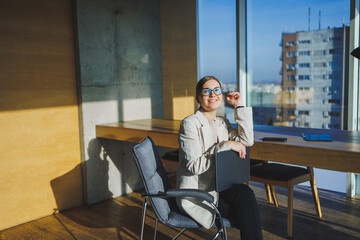 Smiling young businesswoman in casual clothes and glasses sitting in the office with a mobile phone while working on a remote project using a laptop