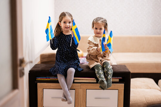 Two sisters are sitting on a couch at home with swedish flags on hands. Sweden children girls with flag .