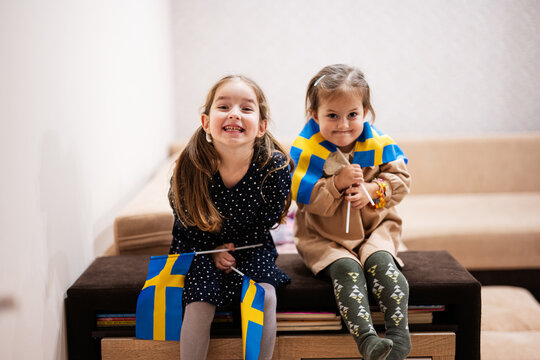Two sisters are sitting on a couch at home with swedish flags on hands. Sweden children girls with flag .