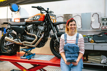 Portrait of smiling mechanic woman holding digital tablet sitting over platform with custom motorcycle on factory