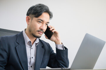 Handsome Asian business man in a suit is sitting and talking on his mobile phone at his desk with a laptop computer.