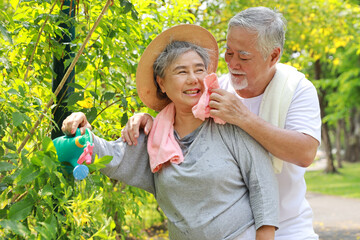 Asian senior couple incasual embracing and spending time in garden backyard while watering flowers plants with laughing and smiling on sunny day after retired. Happy elderly outdoor lifestyle concept.