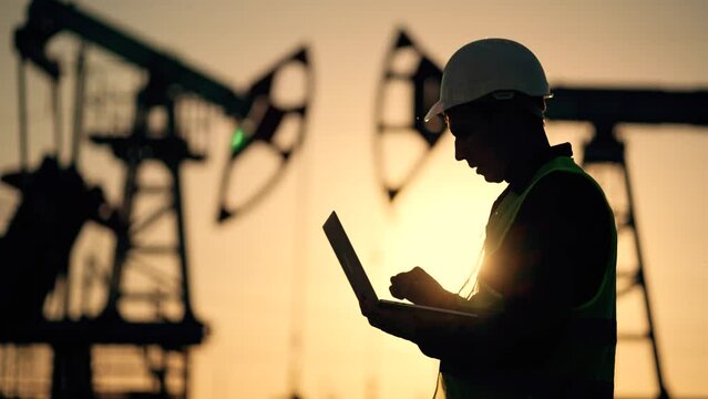 Oil industry. Engineer worker makes notes on operation of an oil pump. The concept of mining. An engineer with laptop and a tablet checks operation of an oil pump. Industrial production of crude oil