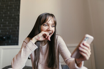 Happy young woman looking at smart phone sitting in bedroom