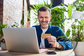 Smiling businessman with laptop holding credit card at loft office
