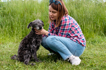 Young woman in pink plaid shirt hugging and petting mixed breed gray fluffy senior dog bedlington...