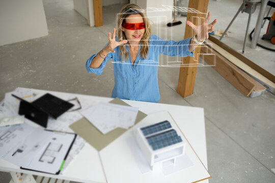 Smiling architect wearing virtual reality simulator gesturing and examining house model