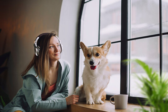 Thoughtful woman wearing headphones sitting with dog near window at home