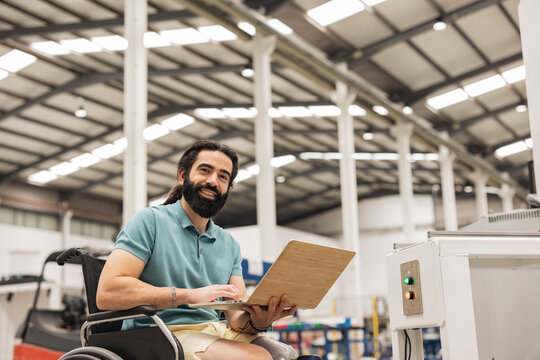 Happy engineer sitting in wheelchair working on laptop in robotics factory