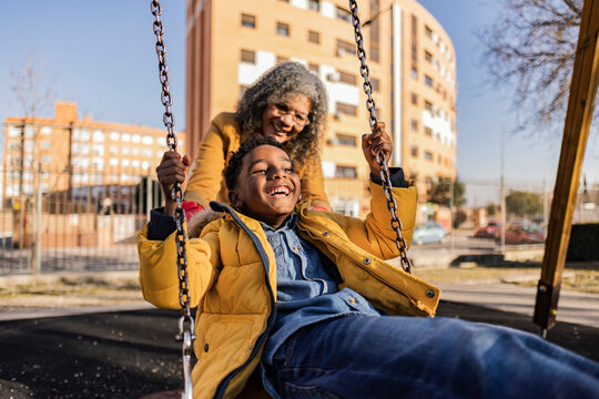 Playful senior woman pushing happy grandson on swing on playground