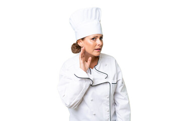 Middle-aged chef woman over isolated background listening to something by putting hand on the ear