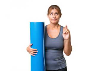 Middle-aged sport woman going to yoga classes while holding a mat over isolated background with fingers crossing and wishing the best