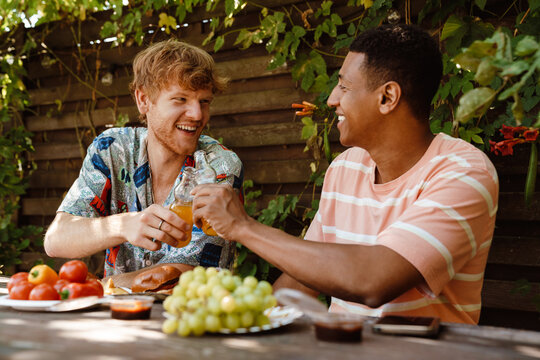 Two cheerful male friends clinking orange soda bottles during barbecue party