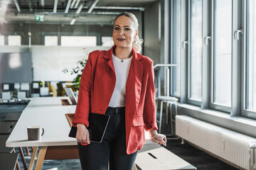 Smiling businesswoman wearing red blazer standing near desk at office